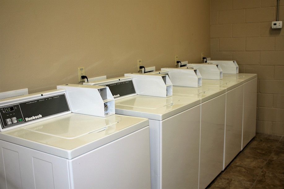 Row of white washing machines in a laundry room, against a tan wall, and a gray-brown floor.