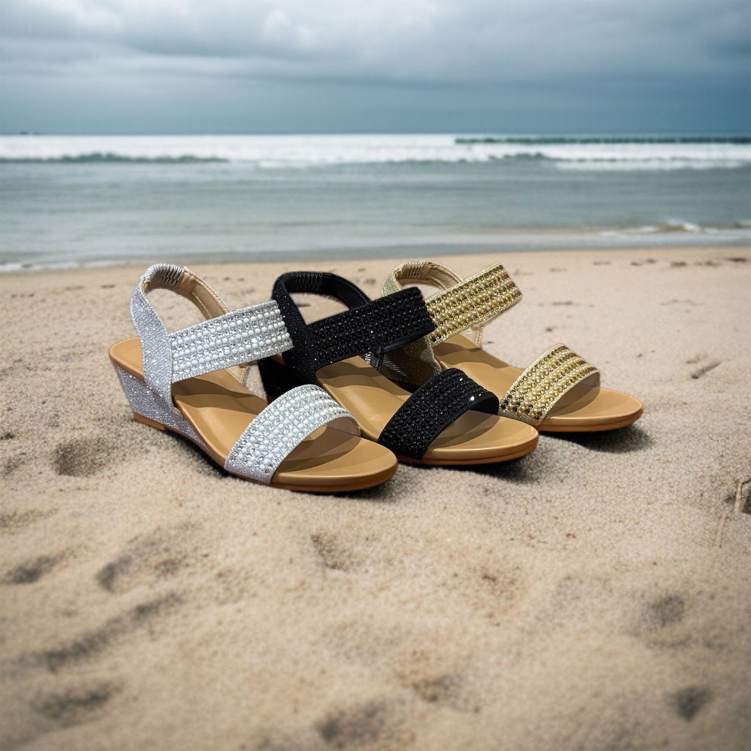 Three embellished sandals on a sandy beach, silver, black, and gold, with the ocean in the background.