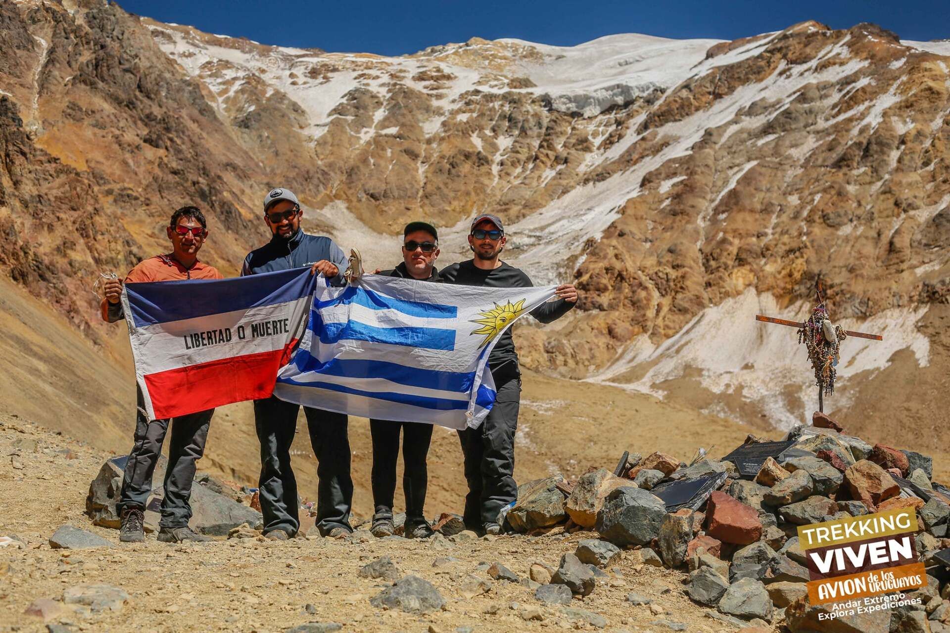 Cuatro excursionistas sostienen banderas en un sendero rocoso de montaña, con picos nevados al fondo.