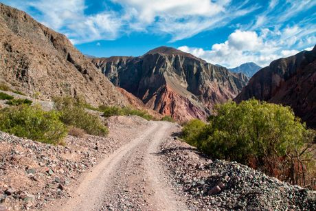 Un camino de tierra pedregoso serpentea entre montañas desérticas de colores bajo un cielo azul
