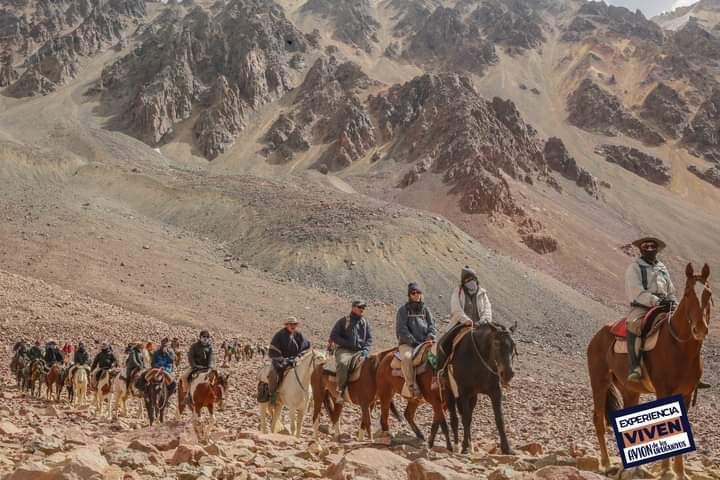 Fila de jinetes a caballo cruzando un paso montañoso rocoso bajo empinados picos áridos