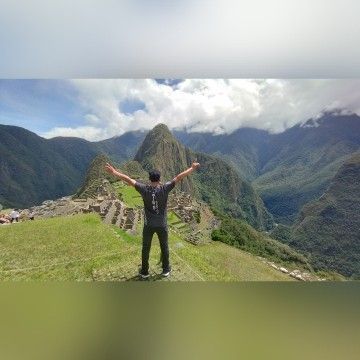 Persona con los brazos en alto en una cresta montañosa cubierta de hierba, con vistas a Machu Picchu.