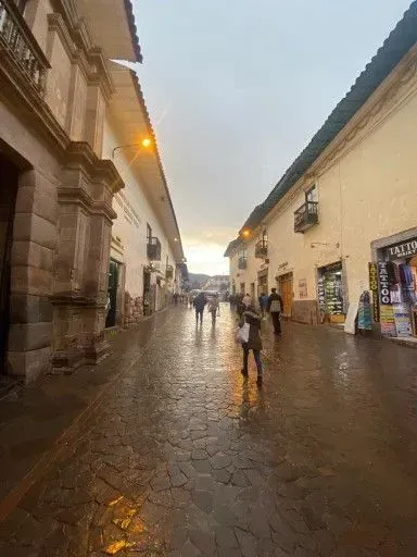 Calle empedrada y lluviosa entre edificios antiguos, con algunos peatones y luces cálidas al anochecer