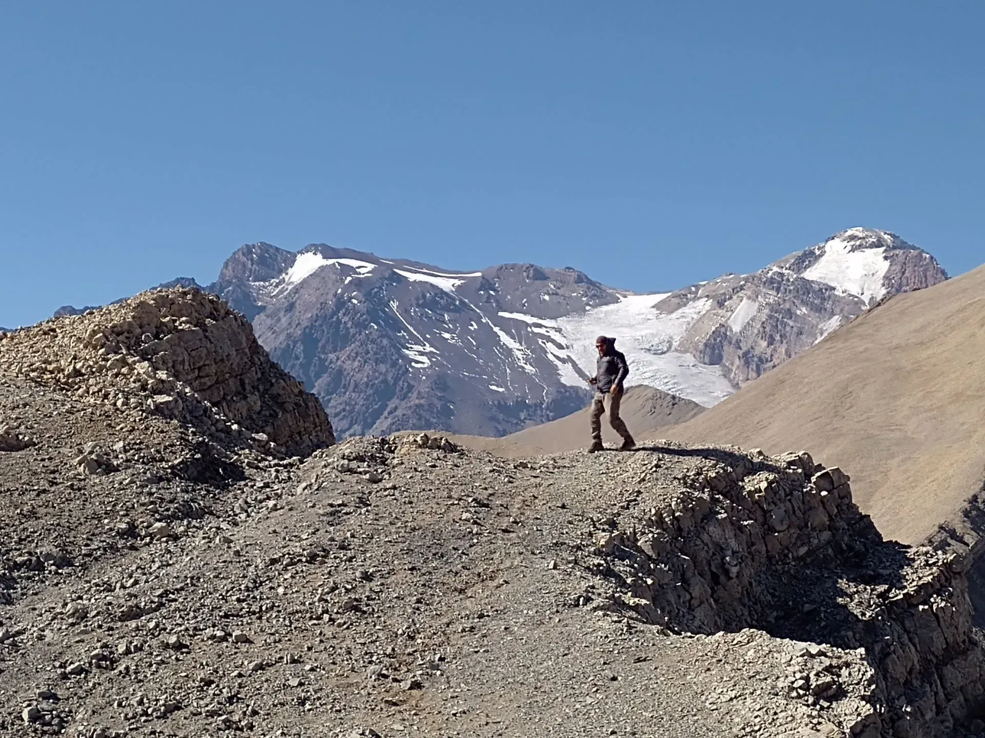 Excursionista de pie en una cresta montañosa rocosa, con picos nevados y cielo azul de fondo