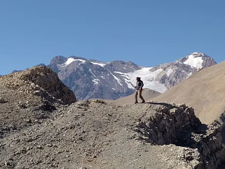 Excursionista de pie en una cresta rocosa de montaña, con picos nevados bajo un cielo azul despejado