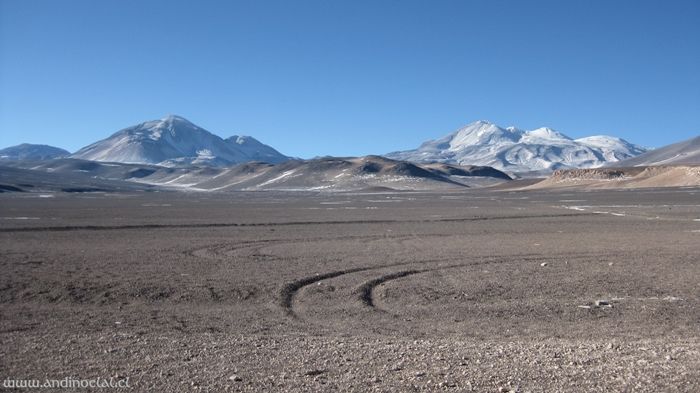 Montañas nevadas se alzan sobre una llanura desolada con huellas de neumáticos bajo un cielo azul despejado