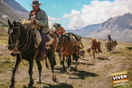 Jinetes a caballo cruzando un valle montañoso cubierto de hierba bajo un cielo azul intenso