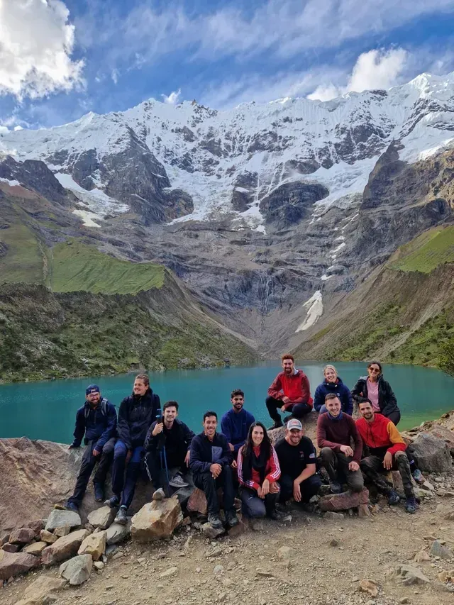 Un grupo de excursionistas posando junto a un lago de montaña de color turquesa, con picos nevados y nubes al fondo