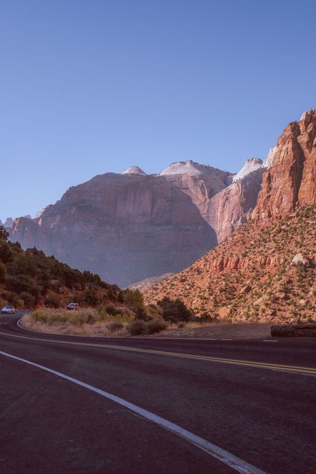 Carretera de montaña con acantilados rojizos y picos nevados bajo un cielo azul despejado.