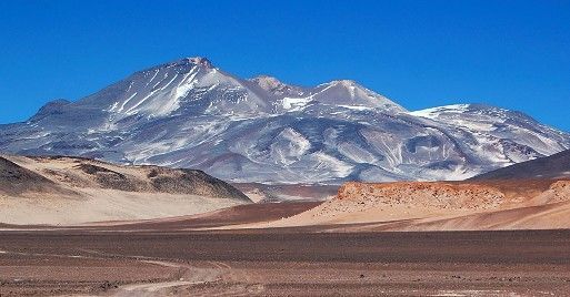 Montaña nevada bajo un cielo azul despejado, con un desierto árido y colinas rojizas en primer plano