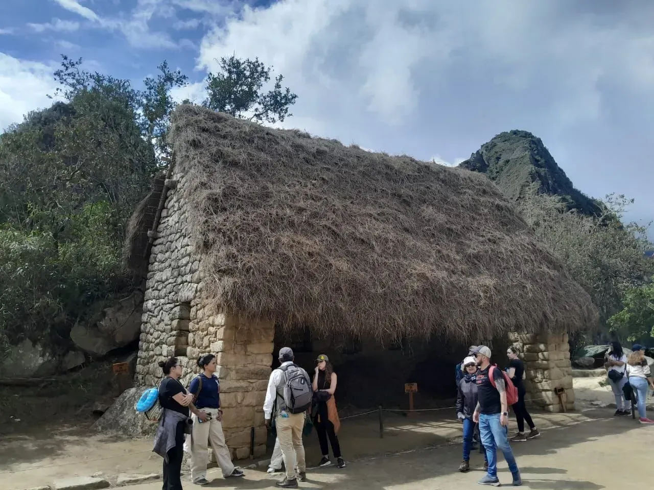 Cabaña de piedra con techo de paja y visitantes en un entorno tropical al aire libre