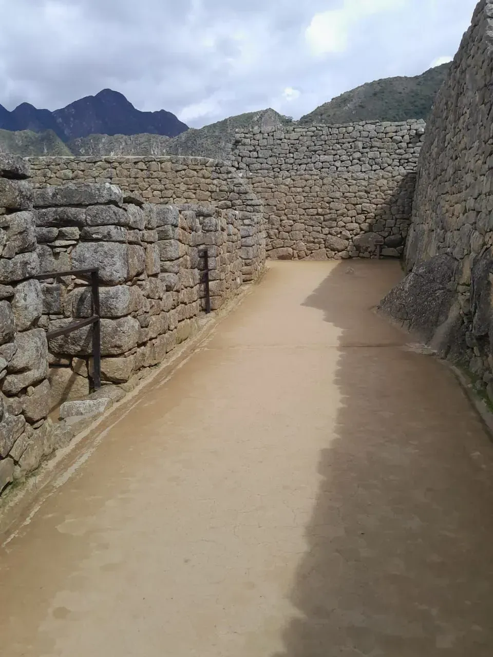 Un sendero de piedra entre antiguos muros de piedra en unas ruinas, con montañas al fondo bajo un cielo despejado.