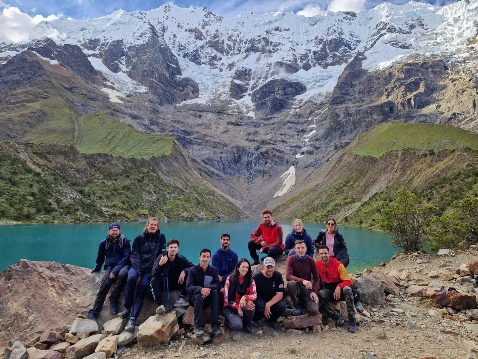 Grupo posando junto a un lago de montaña de color turquesa, con picos nevados y laderas rocosas al fondo
