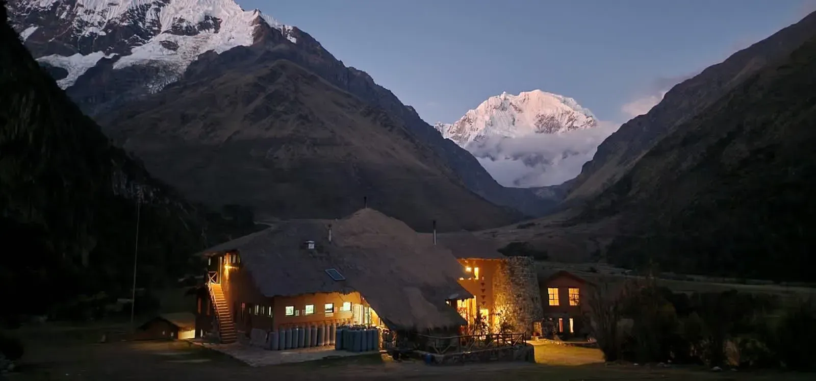 Un refugio de montaña iluminado al anochecer entre valles oscuros, con picos nevados al fondo.