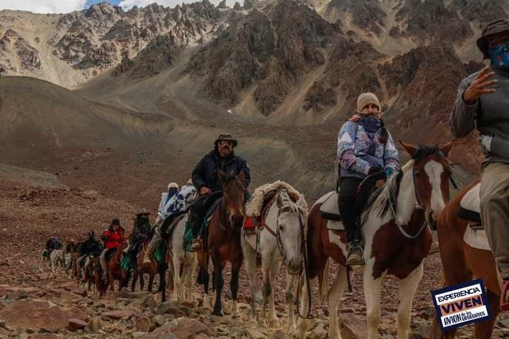 Fila de jinetes a caballo por un sendero rocoso de montaña bajo un cielo nublado