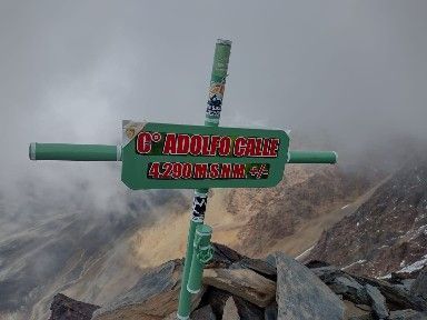 Señal verde de la cima en una cresta rocosa bajo un cielo nublado, con letras rojas y blancas