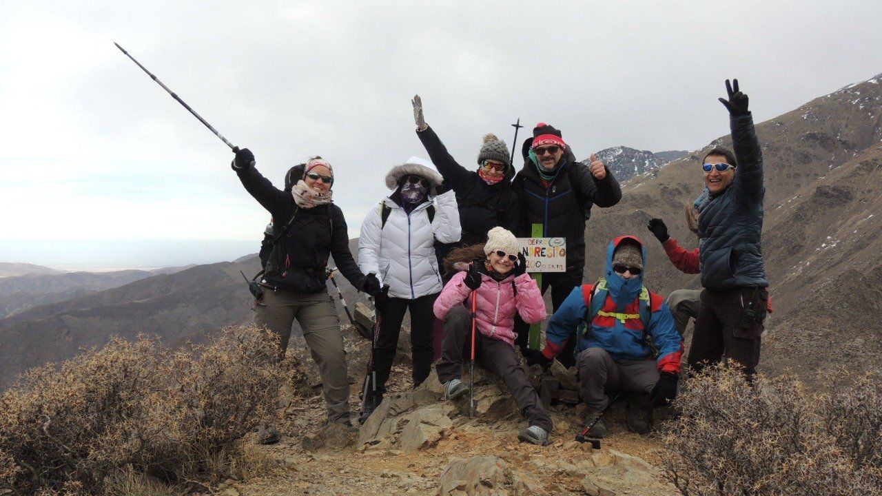 Grupo de excursionistas posando en la cima de una montaña, sonriendo y levantando los brazos bajo un cielo nublado.
