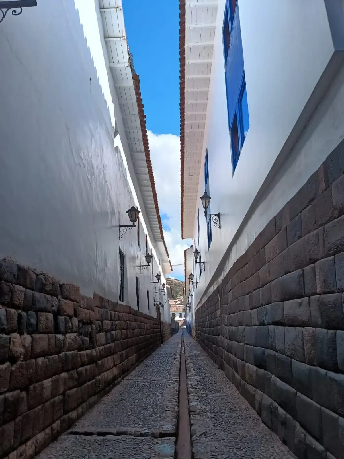 Callejón estrecho de piedra entre muros blancos con detalles azules, que conduce a un cielo azul intenso.