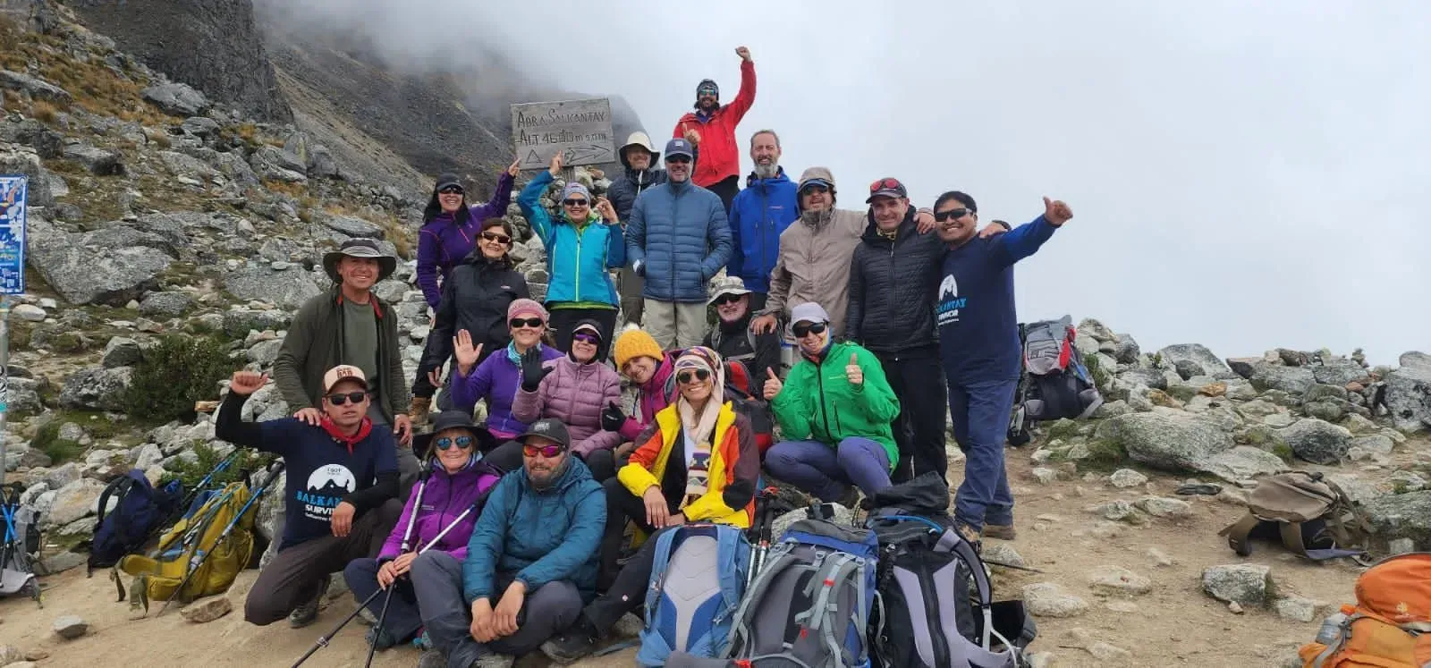 Grupo de excursionistas posando en un sendero rocoso de montaña, algunos sonríen y levantan los brazos bajo un cielo nublado