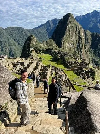Turistas en unos escalones de piedra con vistas a las ruinas de Machu Picchu, con empinadas montañas verdes al fondo