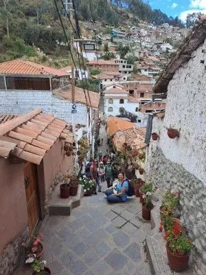 Callejón empedrado en un pueblo de montaña, con tejados de terracota, plantas en macetas y dos personas.