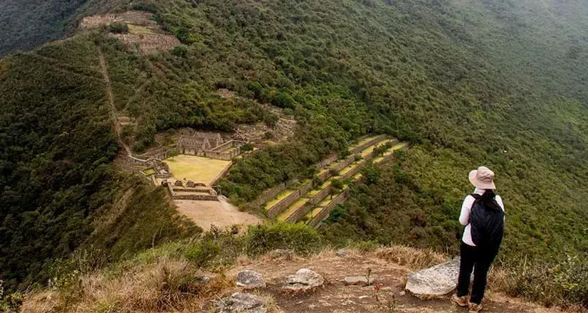 Una persona con sombrero contempla un valle montañoso verde, con antiguas ruinas de piedra debajo.