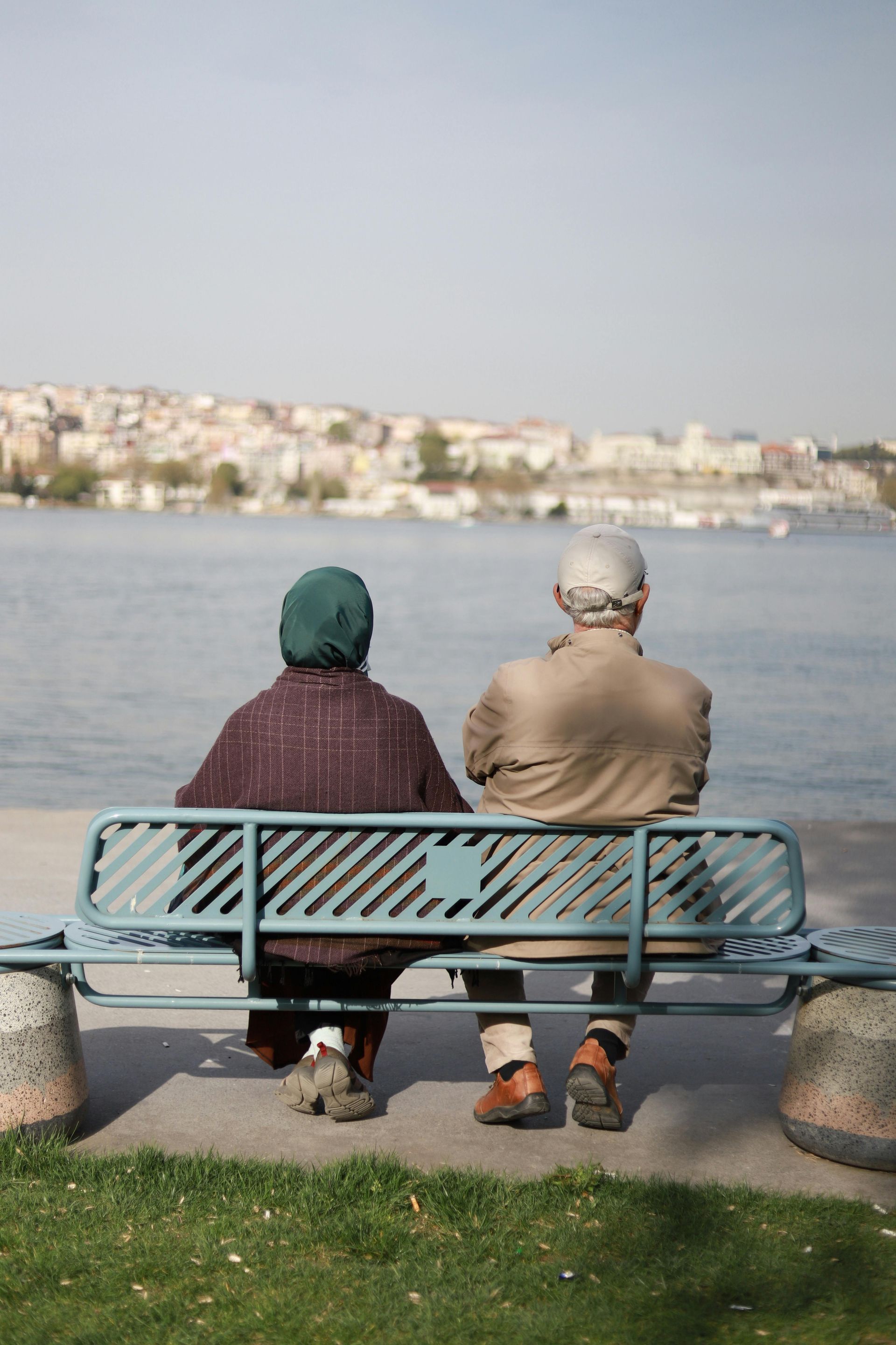 Two people sit on a bench by the water, facing a distant shoreline with buildings.