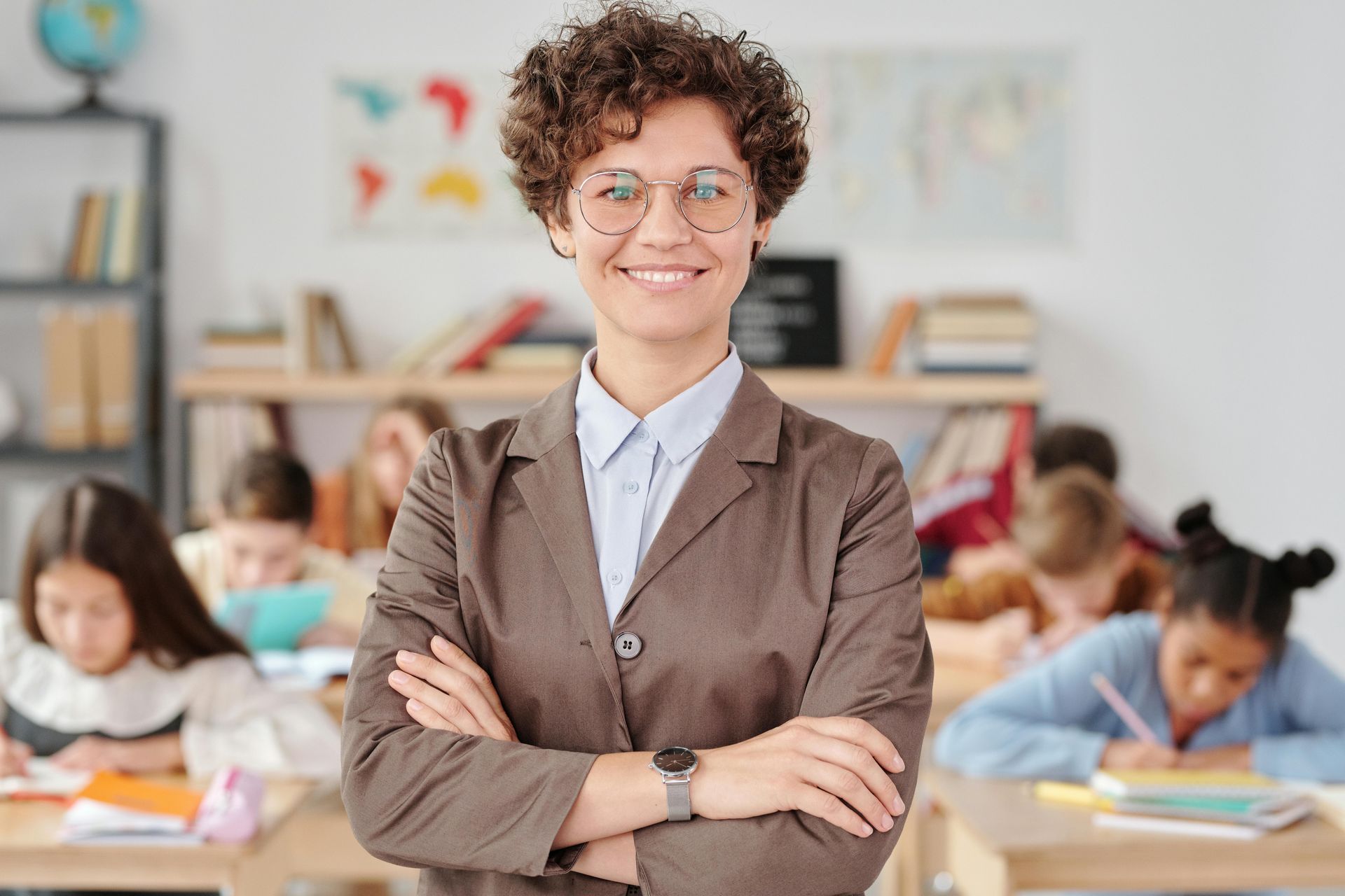 Teacher standing in a classroom with arms crossed, students working in the background.