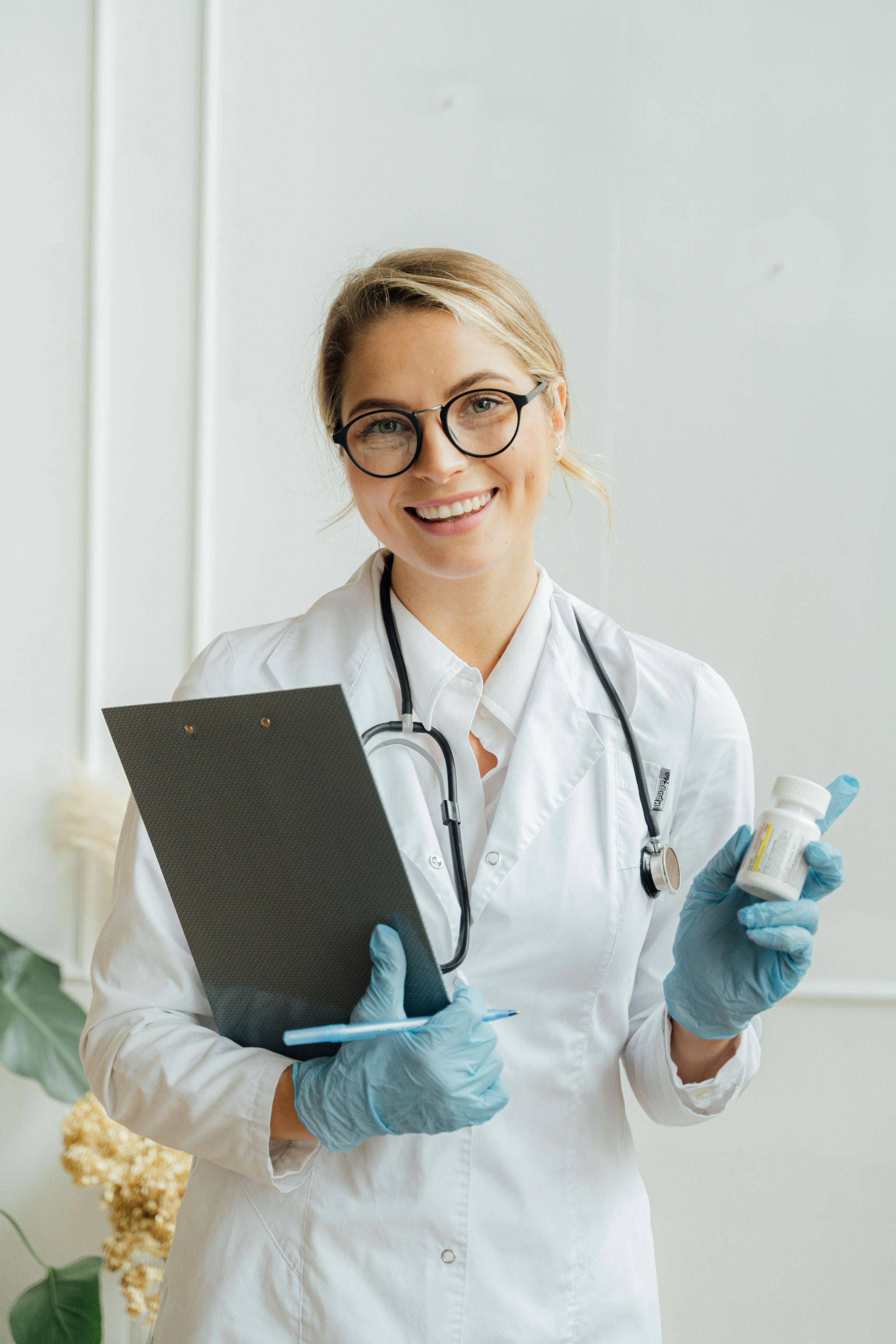 Doctor in white coat holding a clipboard and vial, wearing gloves and smiling in a bright clinic room