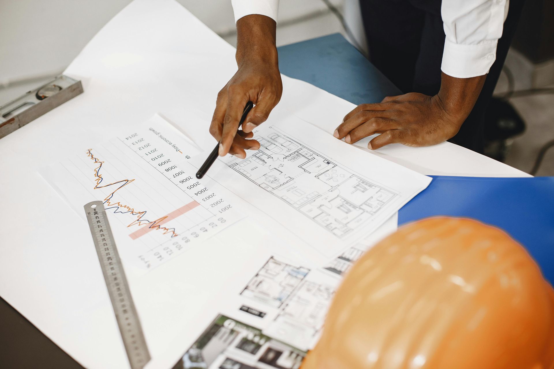 Person pointing at architectural blueprints with a pen, with ruler and orange hard hat on a desk
