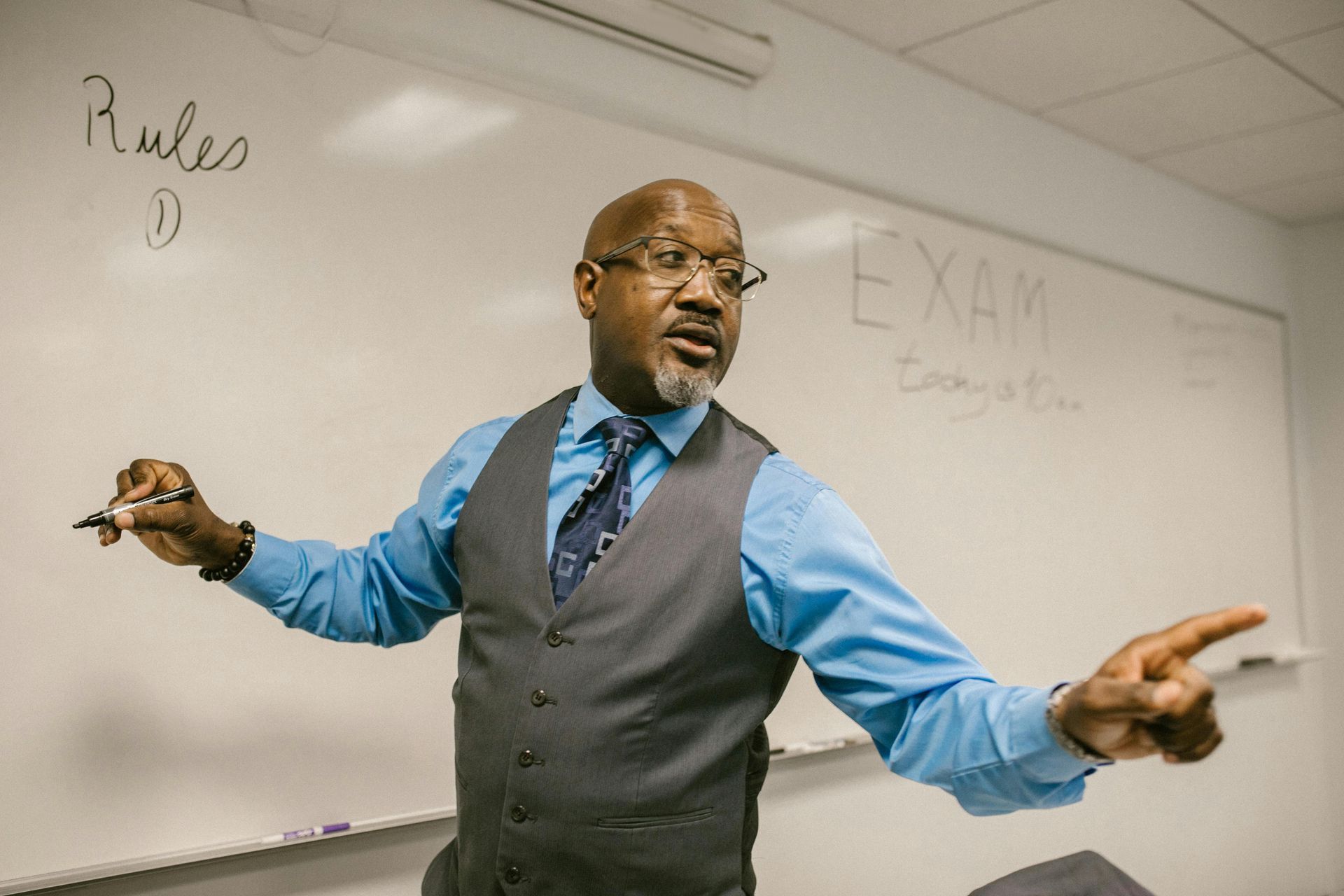 Teacher gesturing in a classroom beside a whiteboard with “EXAM” written on it