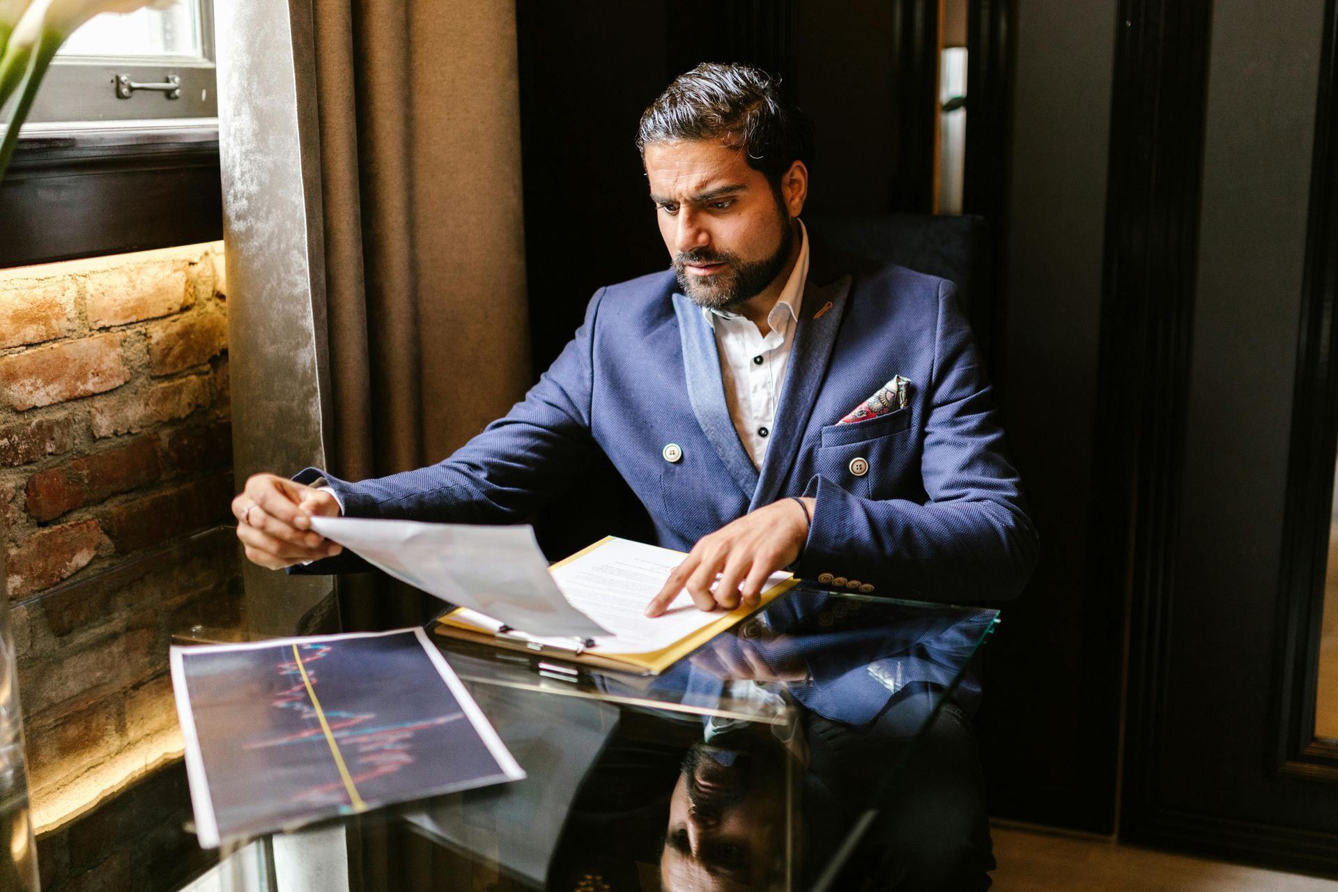 Man in a navy suit reviewing papers at a glass table in a dimly lit office