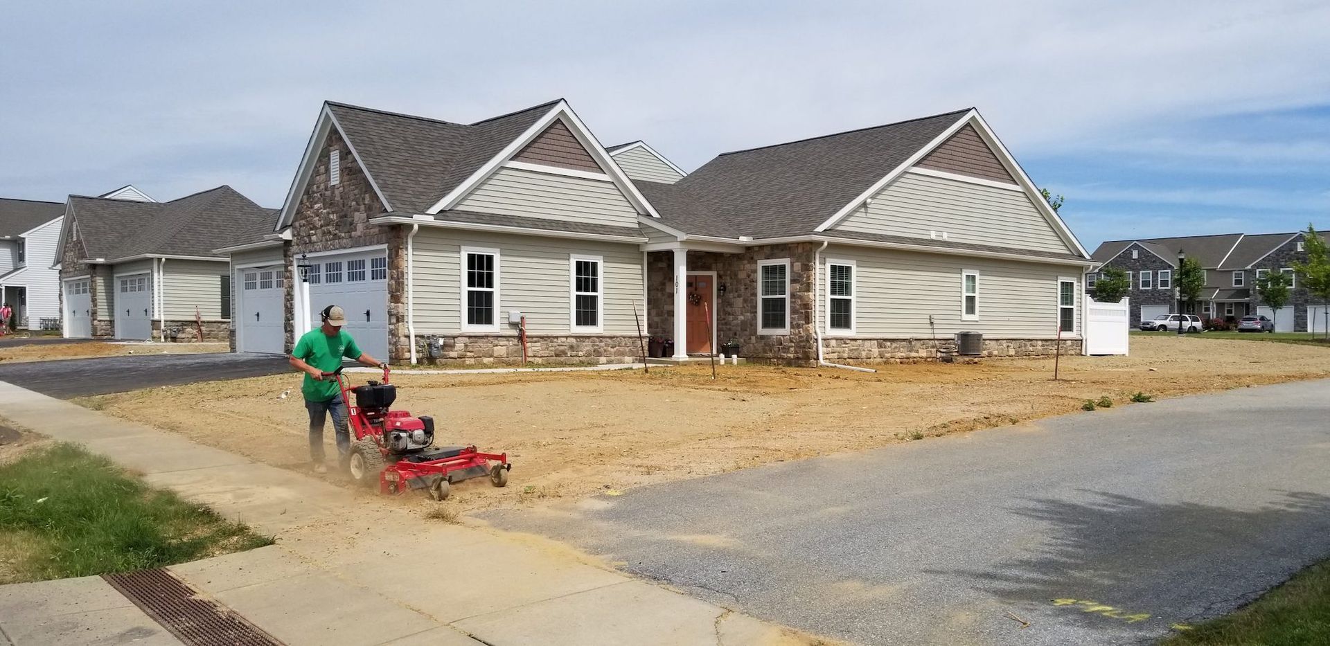 A man is pushing a lawn mower down a dirt road in front of a house.