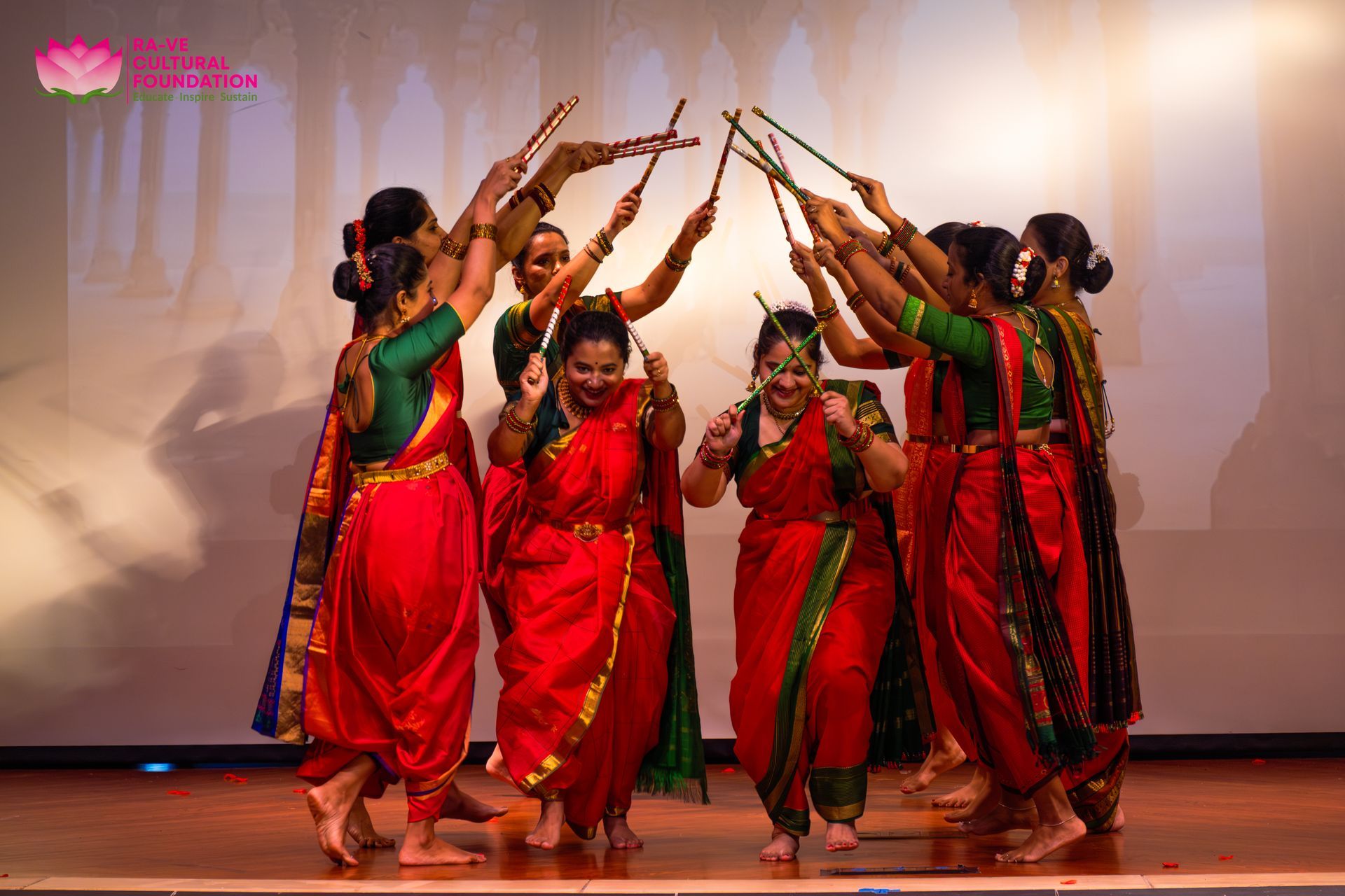 Dancers in red and green costumes perform a synchronized folk dance on stage with raised sticks