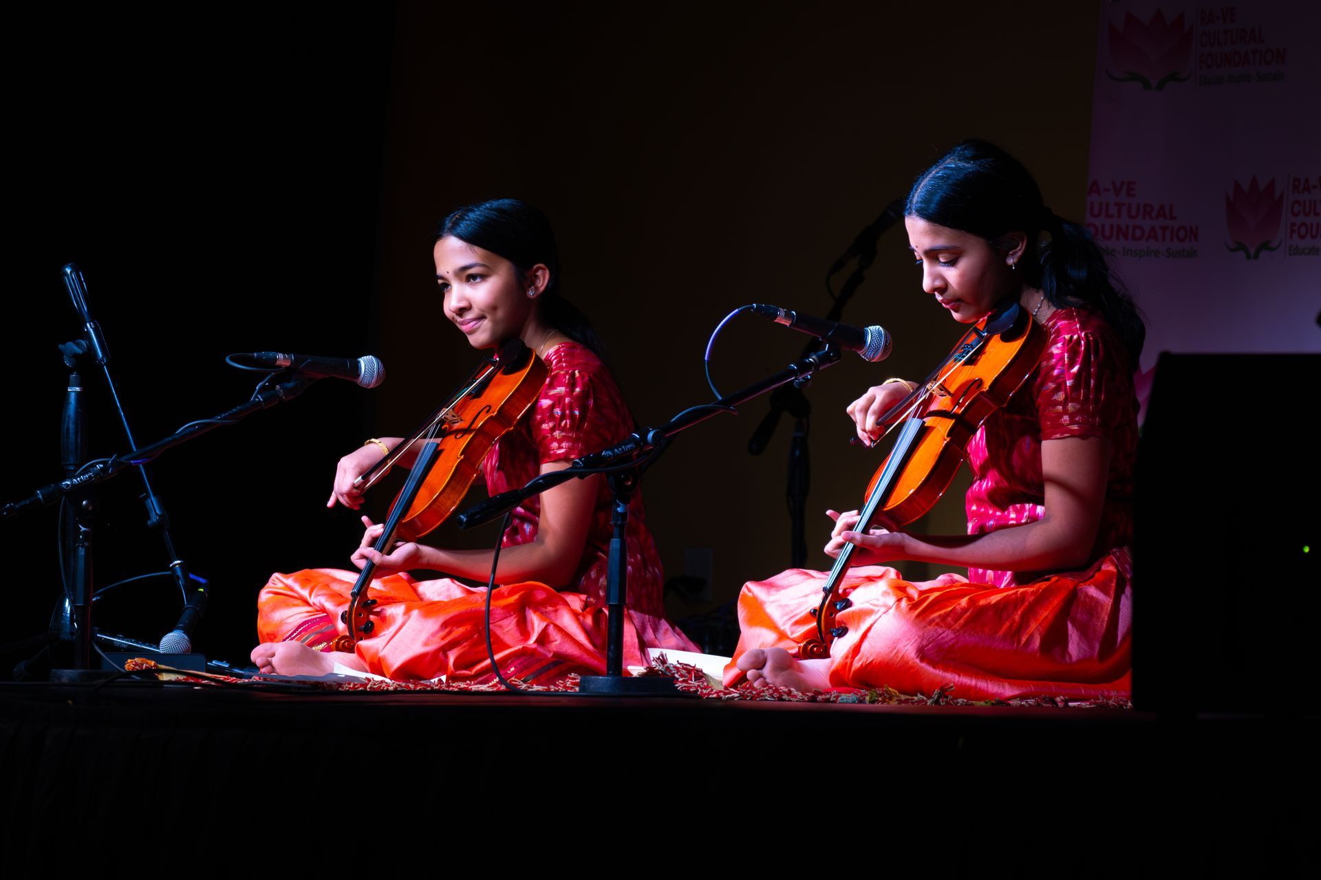 Two women in red sarees playing violins on stage with microphones and warm spotlights