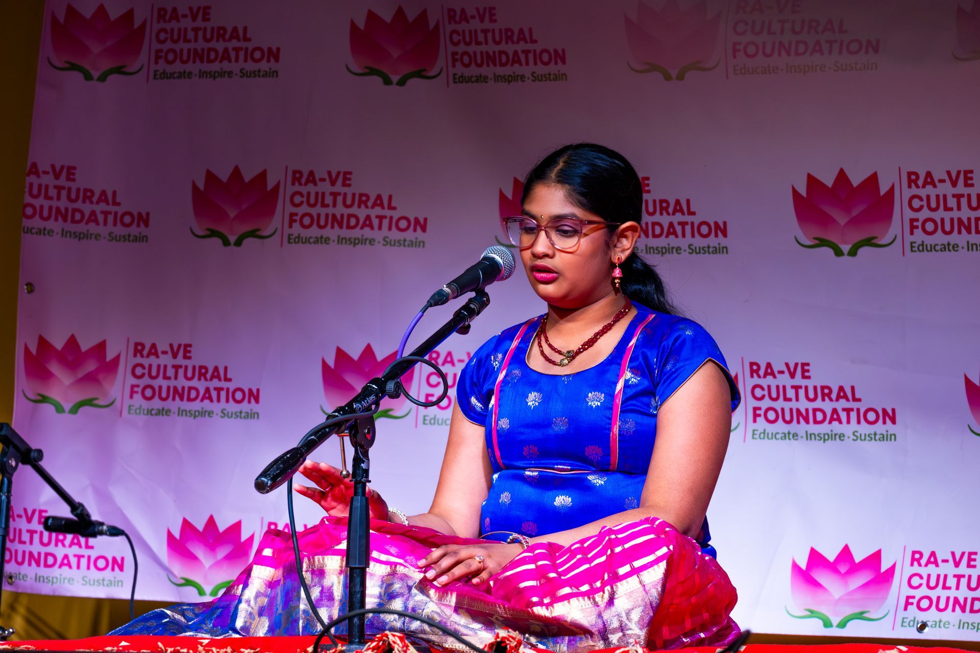 Woman in blue and pink sari speaking at a microphone on a stage with a Ra-Ve Cultural Foundation backdrop