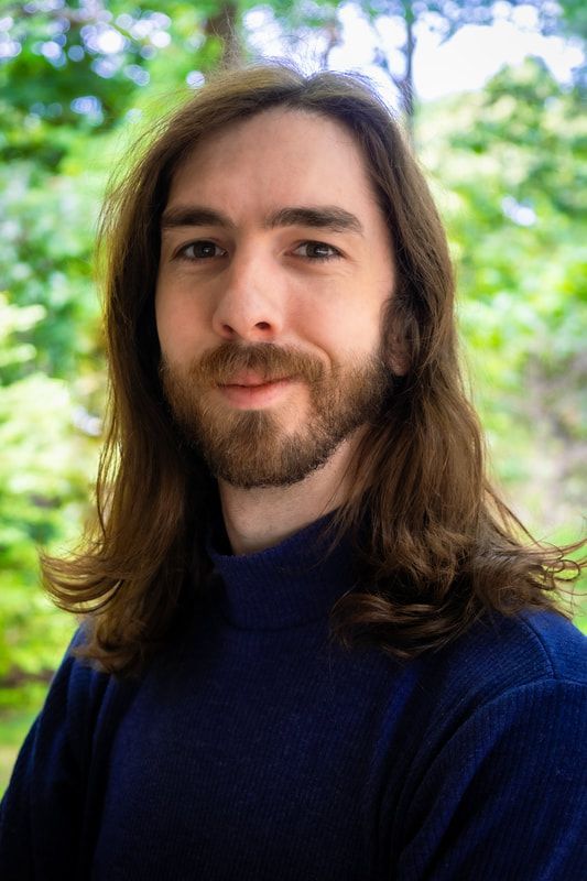 Man with long brown hair and a beard, wearing a blue turtleneck, smiles in a natural outdoor setting.