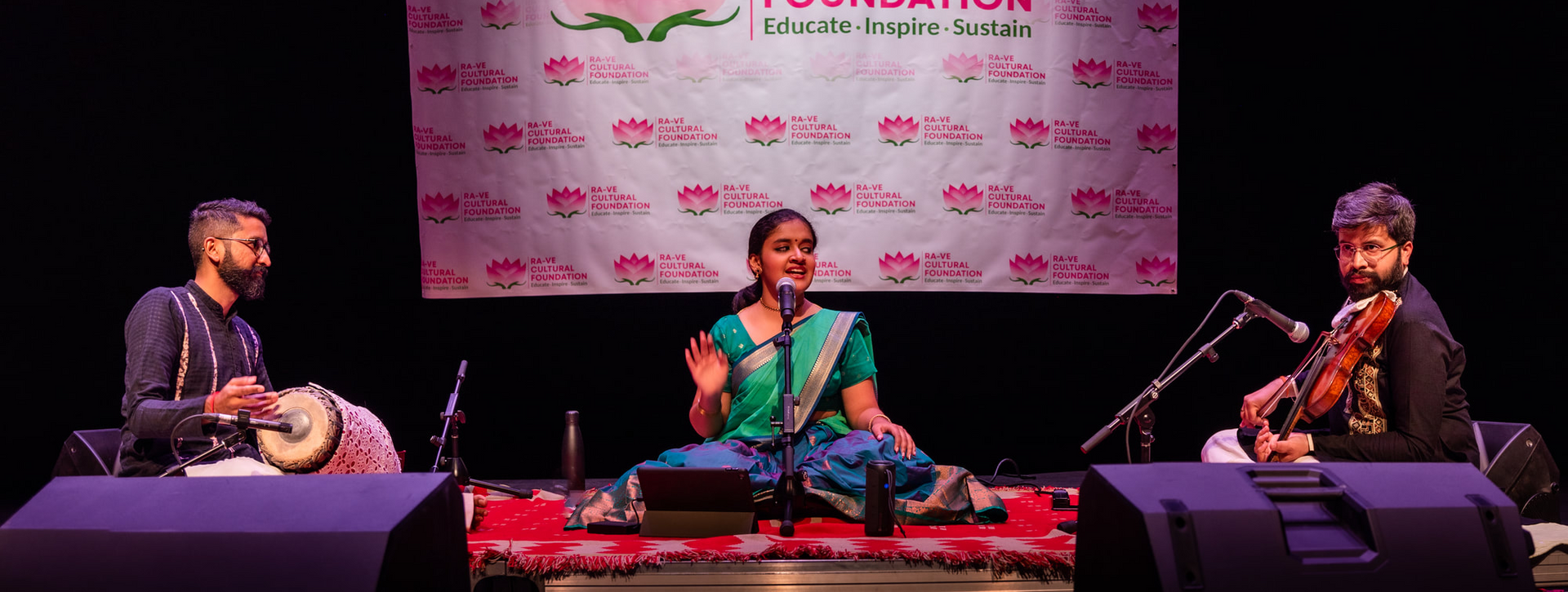 Three musicians perform on a stage; a vocalist with green sari, drummer, and violinist. A banner is in the background.