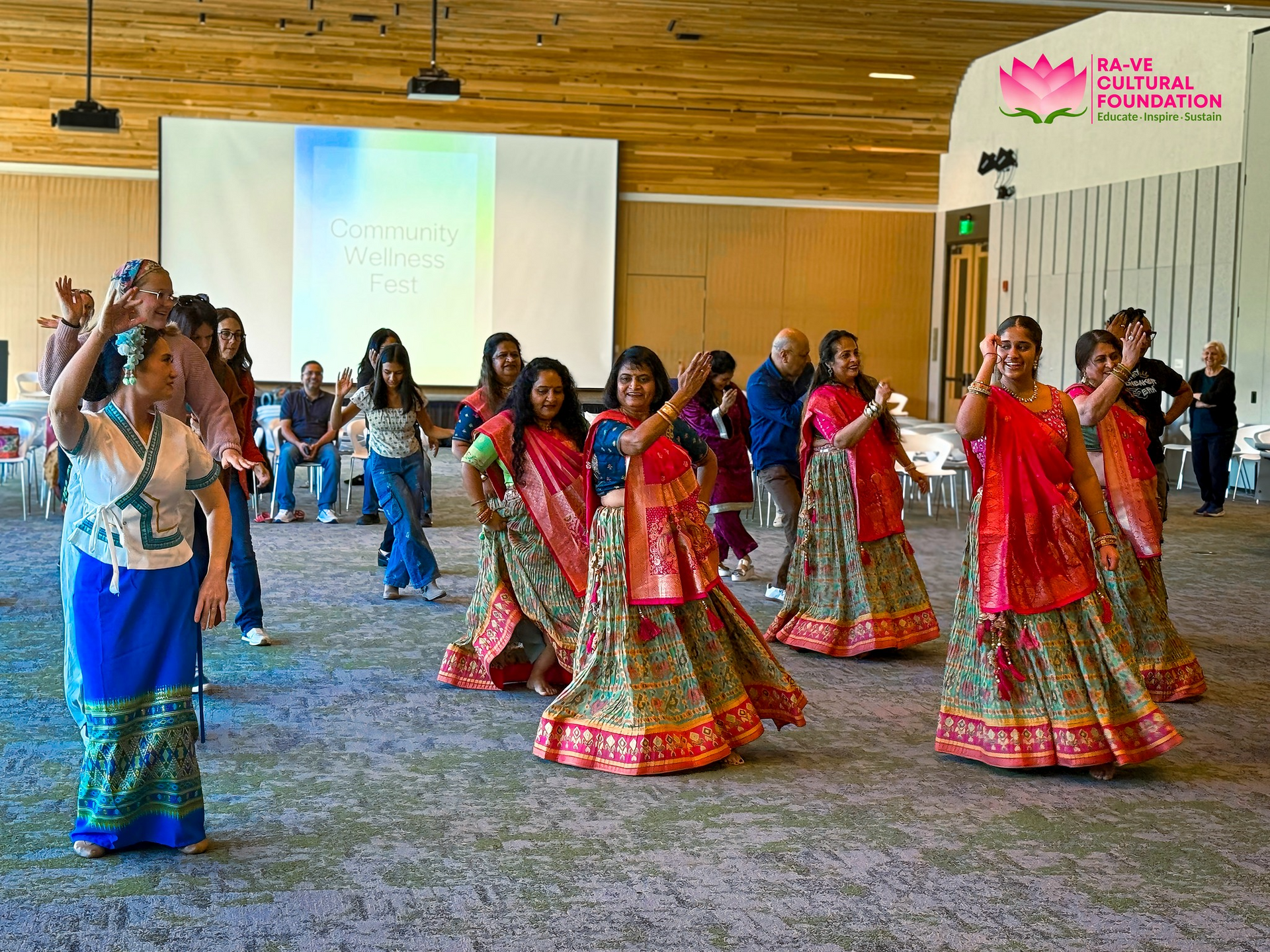 People dancing in colorful saris at an event. Audience watches in background; logo on the right.