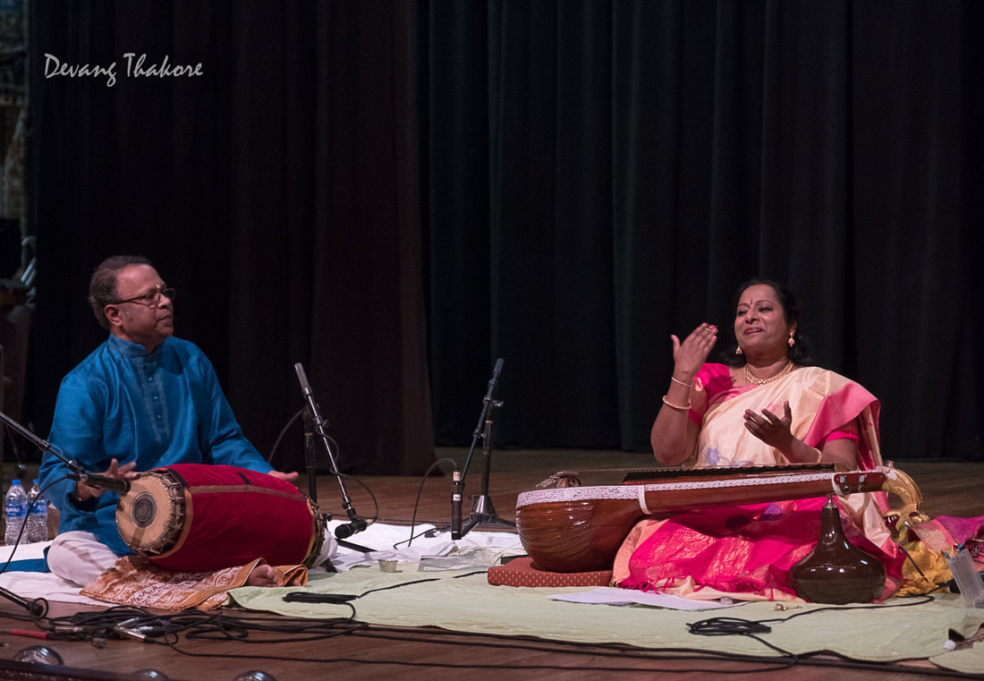 Man plays a percussion instrument, woman sings and plays a stringed instrument on stage.
