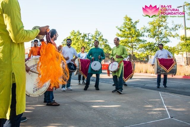 People playing drums in a street. Colorful clothing, green trees, and the logo of