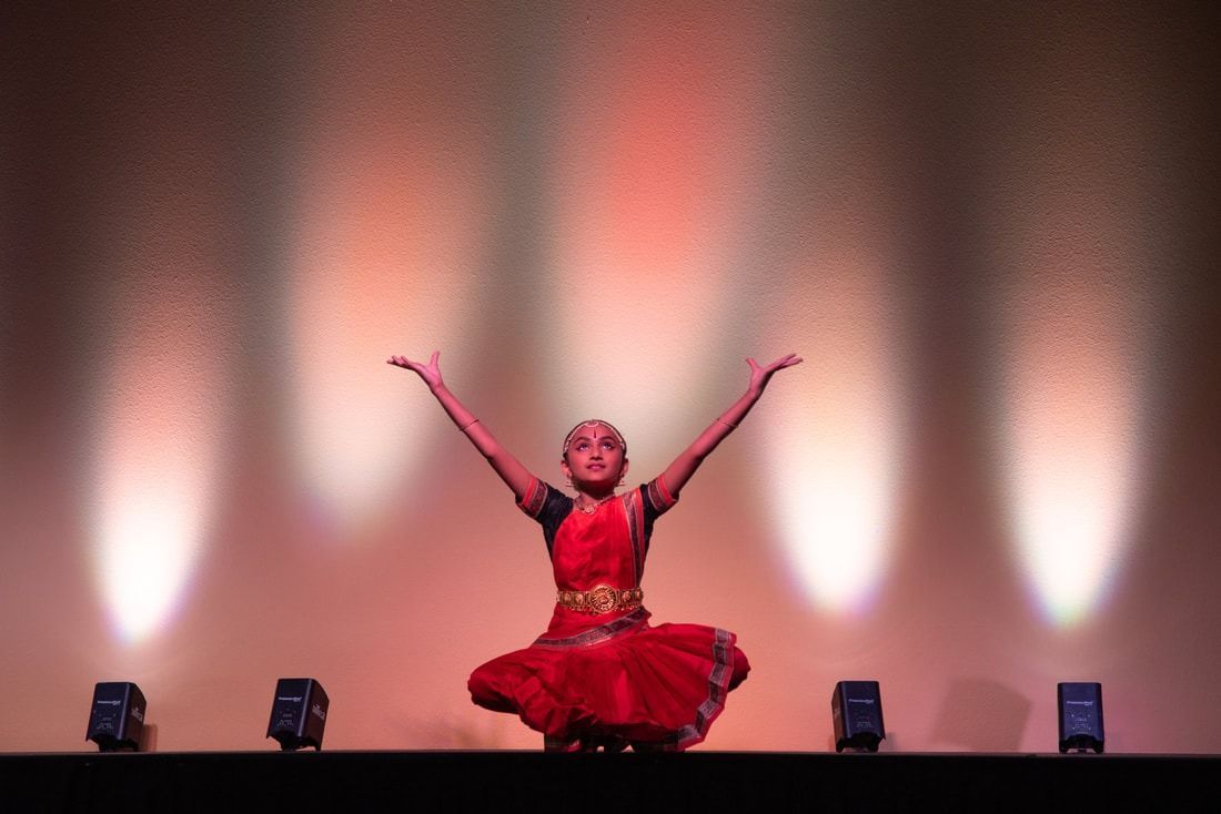 Young dancer in a red costume with arms raised on stage under spotlights.