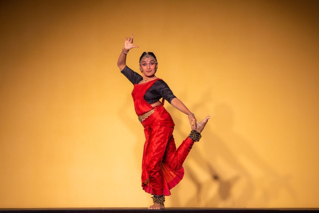 A dancer in red costume, raised arm, bent leg, on stage, yellow backdrop.