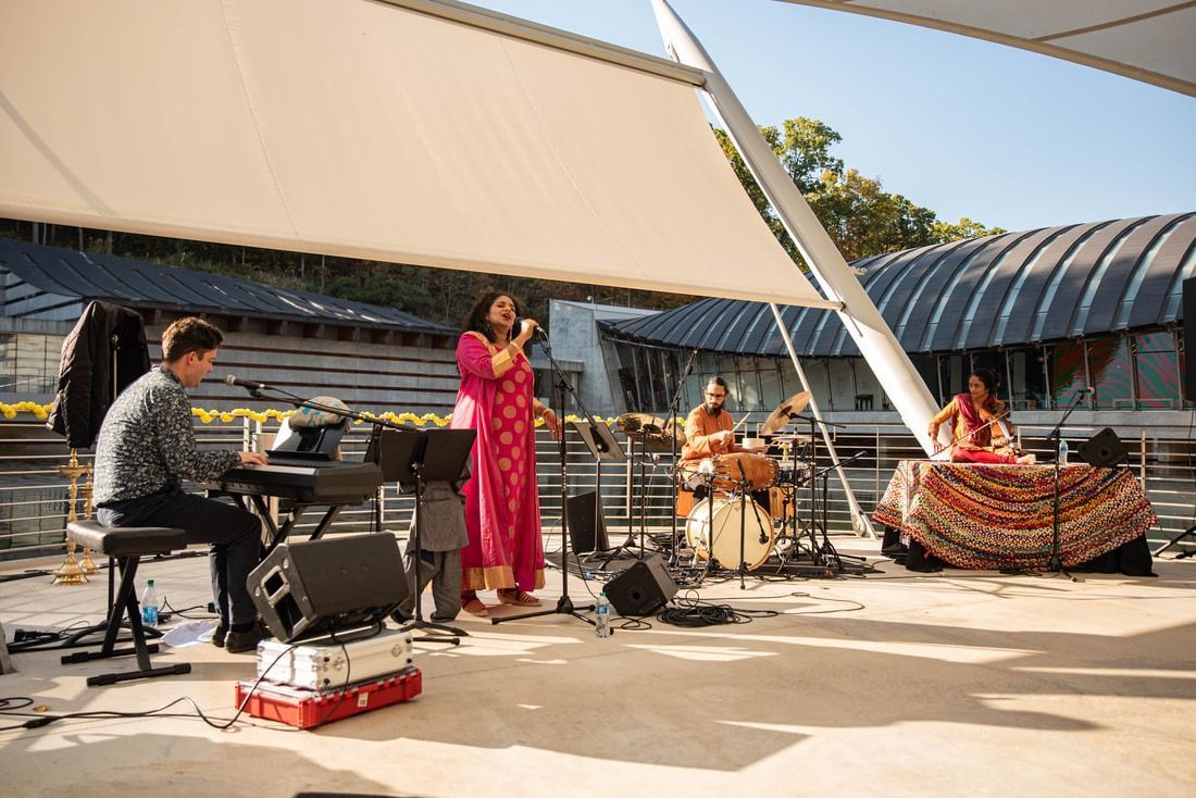 Band performing outdoors under a white canopy. Singer in pink dress, keyboard, drums, and tabla visible.