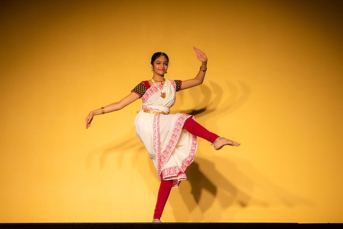 Woman in white and red sari dances on stage with arms raised and leg extended.