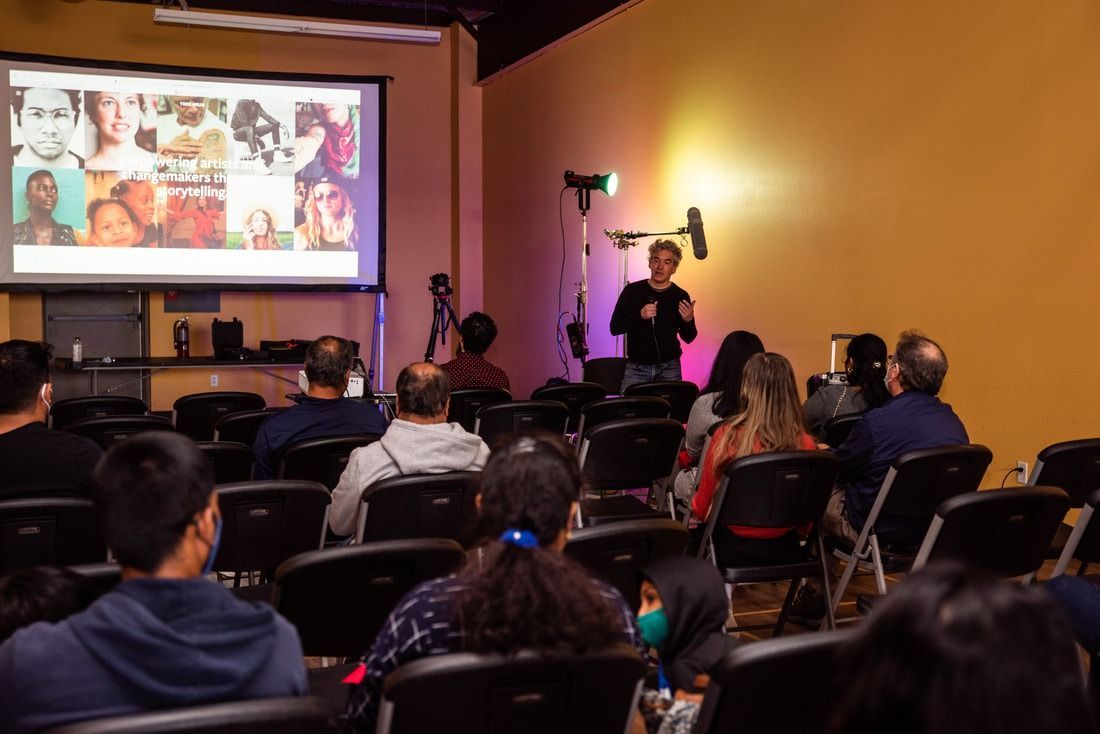 Person presenting to audience in a room; projection screen displays faces.