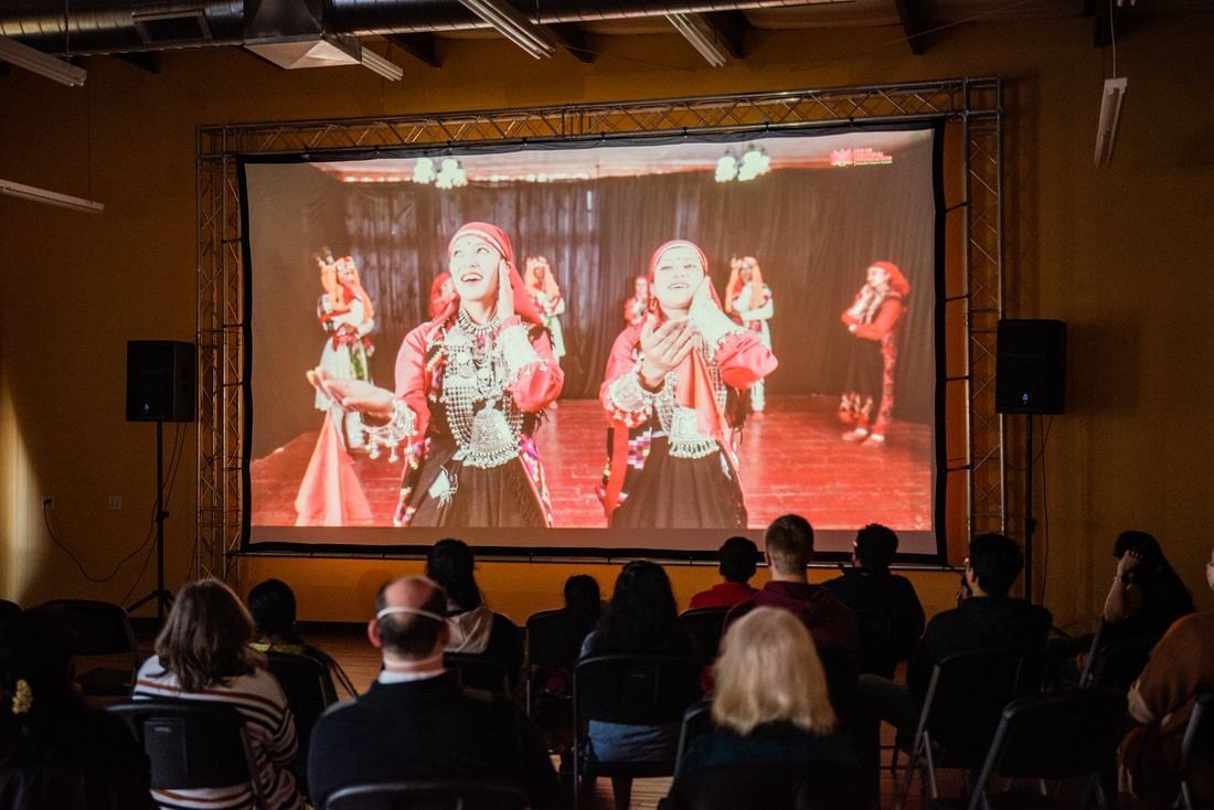 Audience watching a video projection of two people singing onstage. Dark room, stage lighting, and speakers.
