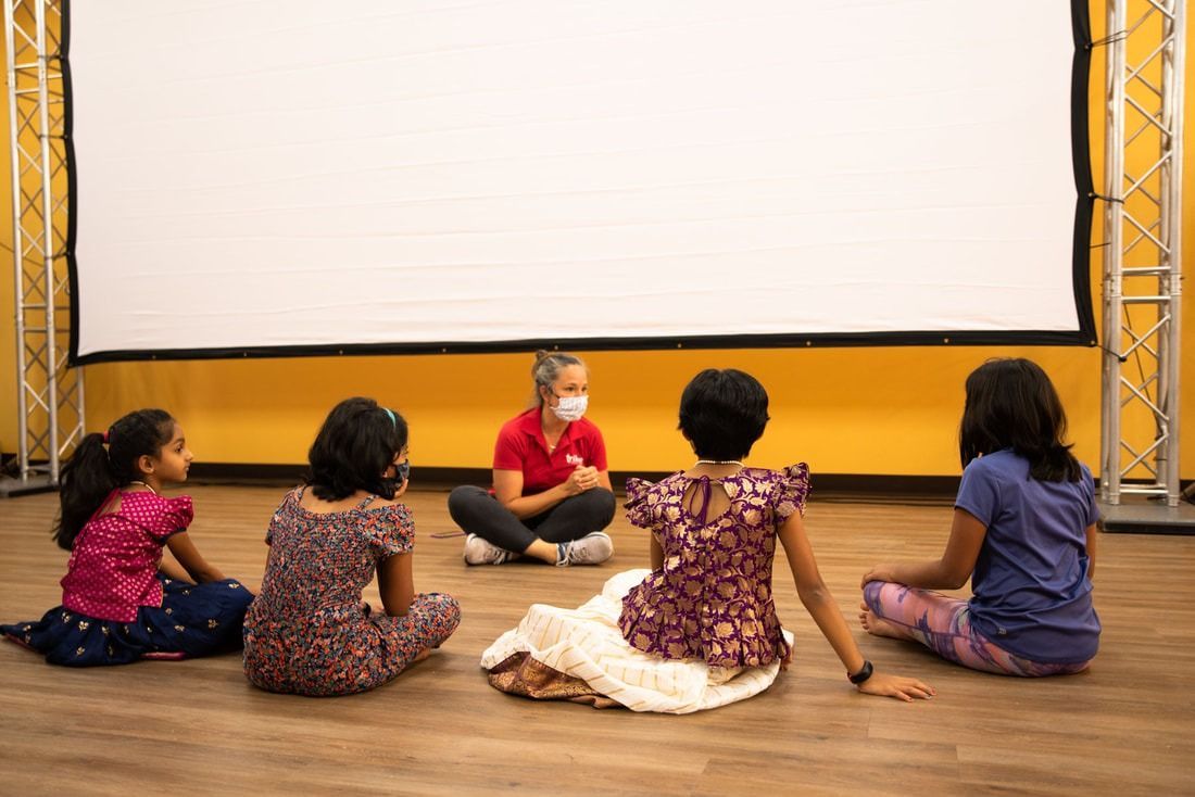 Five children sit on the floor, facing a person wearing a mask, in front of a white screen, and a yellow wall.