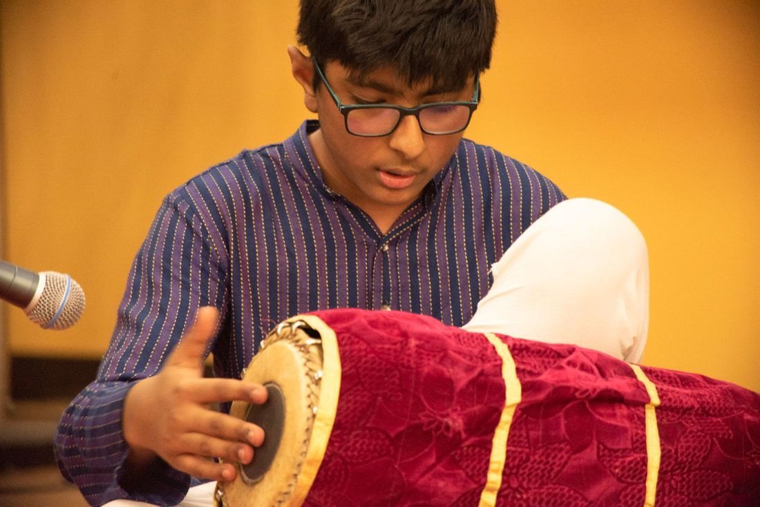 Young person playing a mridangam drum, wearing glasses and a blue shirt, seated with microphone.