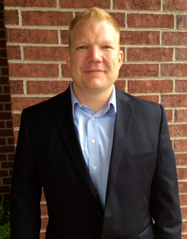 Man in a blue shirt and navy blazer stands in front of a brick wall, smiling slightly.